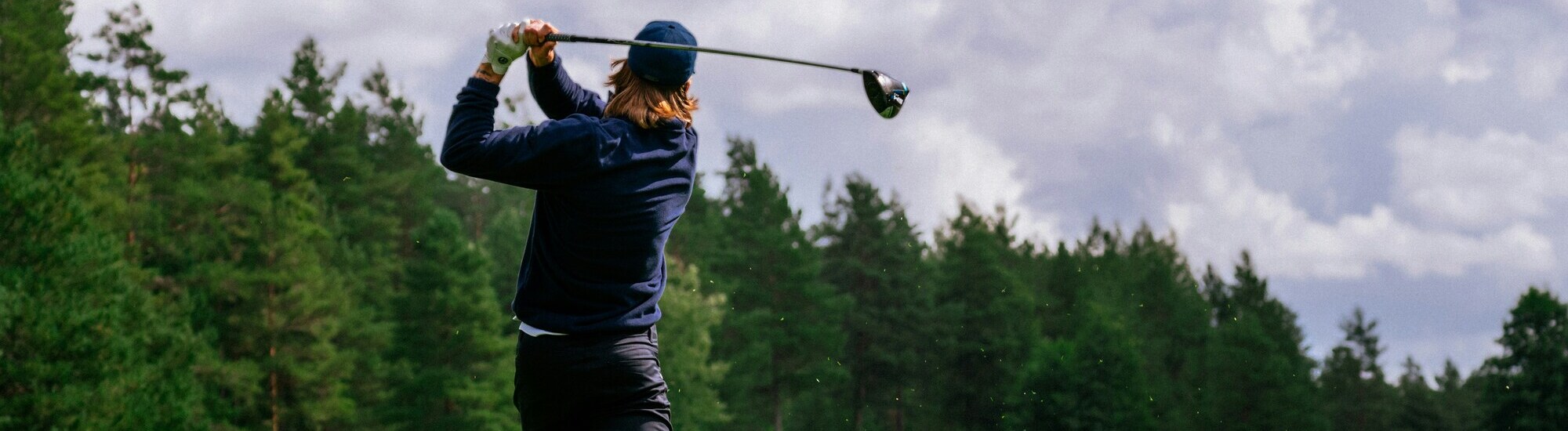 A golfer swings a club on a lush green course under a cloudy sky, surrounded by trees.